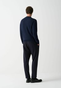 Dunkelblauer Strickpullover mit glatter Textur und geripptem Saum, kombiniert mit lockeren, dunkelblauen Hosen und schwarzen Schuhen, von hinten betrachtet.
