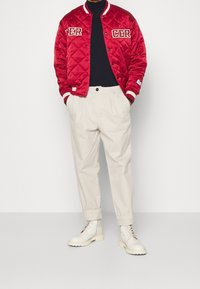 Chaqueta bomber acolchada roja con rayas blancas, parches de logo, camiseta de cuello redondo negra, pantalones beige y botas blancas con cordones.