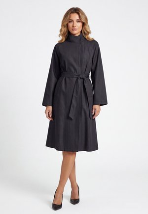 Donna con cappotto nero fino al ginocchio con cintura, maniche lunghe e collo alto, in piedi con le gambe incrociate e tacchi alti neri.