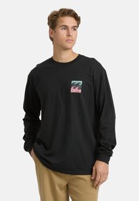 Junger Mann in einem schwarzen Langarmshirt mit mehrfarbigem Wellenmuster und "Billabong"-Schriftzug, kombiniert mit beigen Hosen, der zur Seite schaut.