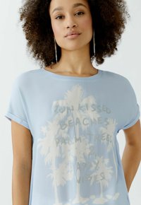 Hellblaues T-Shirt mit kurzen Ärmeln, das eine Grafik von weißen Palmen und den Text "Sonnige Strände, Palmen & Glückliche Vibes" zeigt.