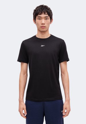Homme debout face à l’avant portant un t-shirt noir Reebok et un pantalon bleu marine sur un fond clair uni.