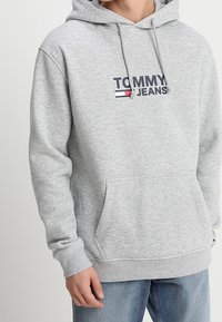 Sudadera de algodón gris con cordones, que presenta un logotipo en navy y rojo de "TOMMY JEANS" en el frente y un bolsillo canguro. Puños y dobladillo acanalados.