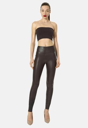 Vrouw met naar achteren gebonden haar, die een zwarte strapless crop top draagt, strakke zwarte leren broek en beige hoge hakken, staand tegen een witte achtergrond.