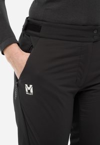 Pantalons softshell noirs au design élégant, dotés d'un logo, de poches zippées et d'une taille ajustable. Le tissu semble lisse avec une finition légèrement mate.