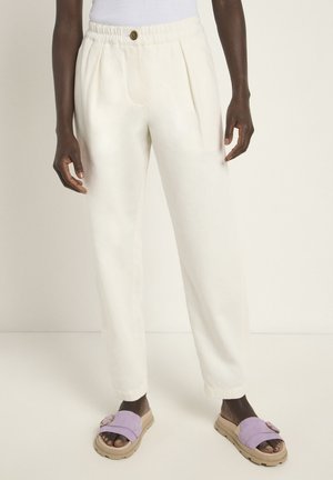Cream-colored, pleated trousers with an elastic waistband and a button detail. Worn with lavender slide sandals featuring a buckle accent.