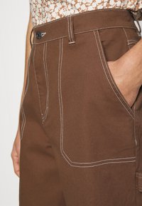 Pantalones marrones con costuras blancas, cierre de botón y bolsillos laterales. La textura de la tela parece resistente y duradera.