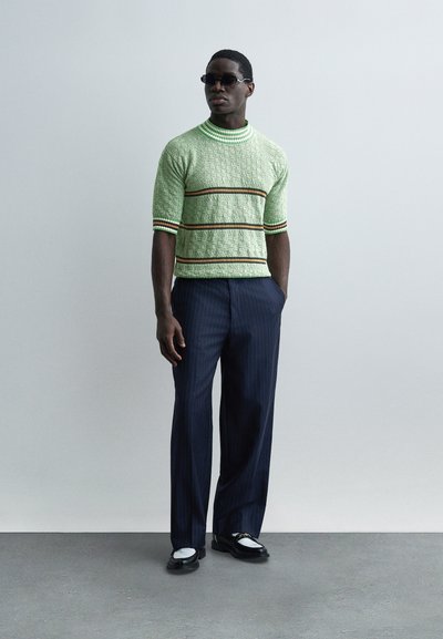 Muž v krátkym rukávom zelenom texturovanom svetri s pruhmi, tmavých nohaviciach s tenkými pruhmi, čierno-bielych topánkach a tmavých slnečných okuliaroch stojaci pri šedej stene.