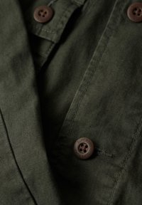 Groene shirt van een textuurstof. Voorzien van bruine knopen, waarvan er één is gelabeld met "SUPERDRY", en een borstzak met stikdetails.