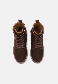 Hnědé semišové kotníkové boty se stejnobarevnými tkaničkami, polstrovaným límcem, vnitřní vložkou a kontrastním prošíváním podél podrážky. Přední zipové zapínání.