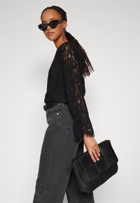 Black lace top with long sleeves, paired with gray jeans. The model holds a black textured handbag and wears black sunglasses.