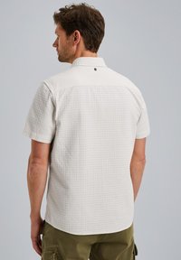 Camisa de botones de manga corta en tejido texturizado color blanco roto con un patrón de cuadrícula, que presenta un collar y un pequeño detalle de logo en la parte posterior.