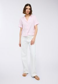 Camicia rosa a maniche corte con colletto, decorata con righe verticali e bottoni frontali, abbinata a pantaloni bianchi a gamba larga e infradito beige.