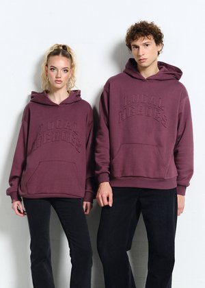 Dwóch młodych dorosłych stoi obok siebie, ubranych w pasujące bordowe bluzy z kapturem z napisem "LOCAL HEROES" oraz czarne spodnie, na jednolitym białym tle.