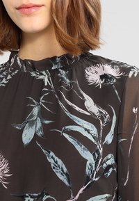 Dunkle Blumenbluse mit gerafftem Ausschnitt, helle botanische Muster in sanftem Blau und Rosa auf tiefbraunem Hintergrund.