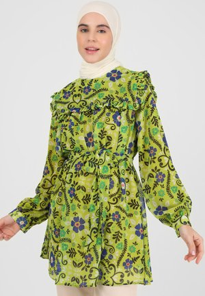 Green patterned tunic with ruffles, featuring floral designs in blue and purple. Long sleeves, belted waist, and lightweight fabric.
