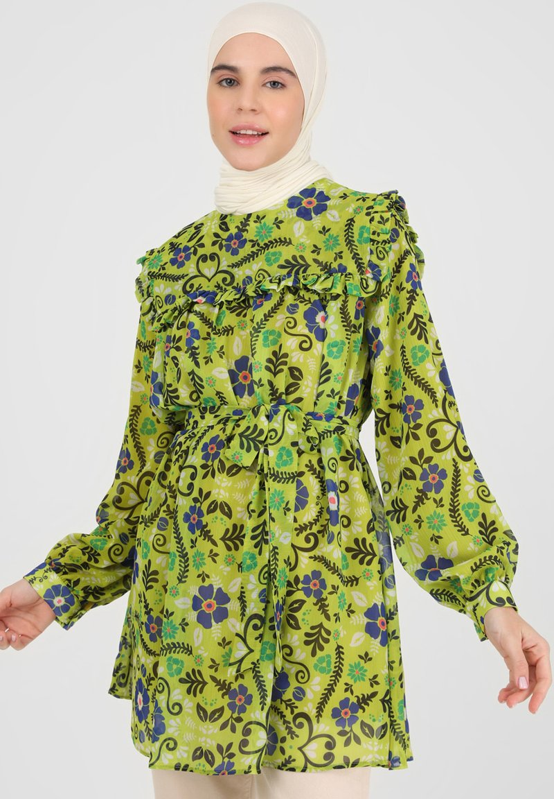 Green patterned tunic with ruffles, featuring floral designs in blue and purple. Long sleeves, belted waist, and lightweight fabric.
