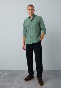 Camicia verde con bottoni e tasca, maniche arrotolate, abbinata a pantaloni scuri e scarpe di camoscio marroni, su uno sfondo chiaro.