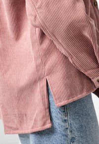 Gros plan d'une personne portant une chemise en velours côtelé rose à côtes avec une fente latérale sur un jean en denim bleu clair.