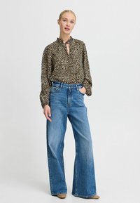 Blusa con stampa leopardata e colletto arricciato, abbinata a jeans blu a vita alta e gamba larga e scarpe beige, su uno sfondo semplice.