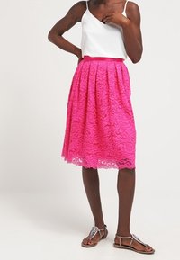 Kobieta ubrana w białą bluzkę bez rękawów, jasnoróżową koronkową spódnicę do kolan i srebrne płaskie sandały, stojąca na jednolitym tle.