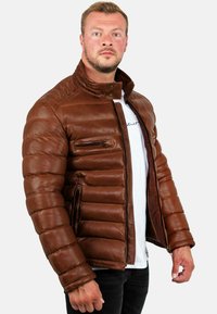 Bruine leren pufferjack met een hoge kraag, ritssluitingen en een horizontaal gestikt patroon. Voorzien van een rits aan de voorkant.