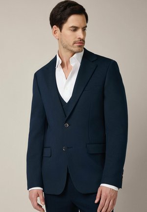 Mężczyzna w ciemnogranatowej marynarce i pasującym kamizelce, na białej koszuli, patrzący w lewo na tle beżowym.