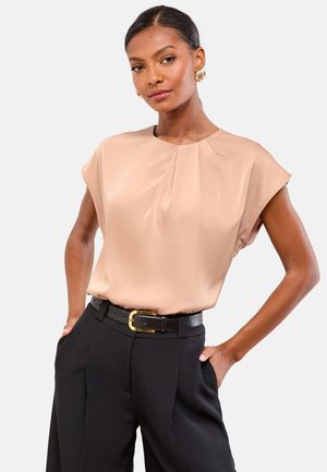 Mujer de pie con las manos en los bolsillos, vistiendo blusa beige de manga corta, pantalones negros y cinturón negro con hebilla dorada, fondo neutro.