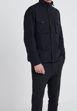 Homme portant une veste noire boutonnée avec quatre poches à rabat sur le devant et un pantalon noir assorti sur un fond uni.