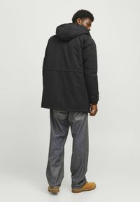 Schwarze Kapuzenparka mit lässiger Passform, ausgestattet mit einer glatten Textur, mittellanger Länge bis zur Oberschenkelmitte und Seitentaschen. Kombiniert mit lockeren grauen Jeans und braunen Stiefeln.