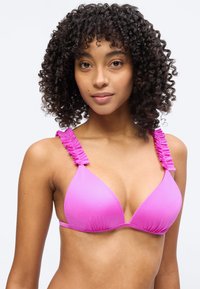 Γυναίκα με σγουρά μαλλιά που φοράει ένα φωτεινό ροζ μπικίνι τοπ με βολάν στα τιραντάκια, κοιτάζοντας μπροστά με φόντο έναν απλό φωτεινό τοίχο.