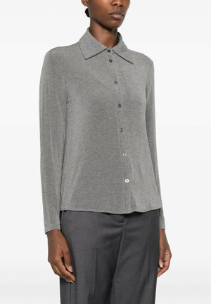Donna che indossa una camicia grigia a maniche lunghe con colletto ampio e pantaloni sartoriali grigio scuro, in piedi davanti a uno sfondo semplice.