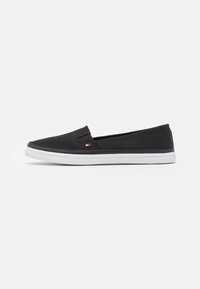 Schwarzer Slip-On Sneaker mit weißer Gummisohle und kleinem rechteckigen rot-weiß-blauen Logo an der Seite, im Profil gezeigt.