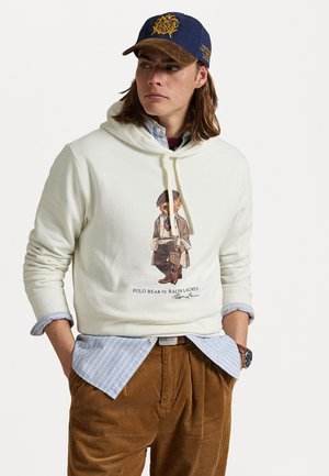 Felpa chiara con cappuccio e design del polo bear stampato, indossata sopra una camicia a righe blu. Combinata con pantaloni in corduroy marroni e un cappellino blu.