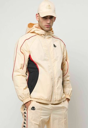 Beżowa kurtka muszelkowa z czarnymi i czerwonymi akcentami, z zamkiem błyskawicznym z przodu, elastycznymi mankietami i kapturem. W komplecie spodnie o dopasowanym stylu z detalami logo.