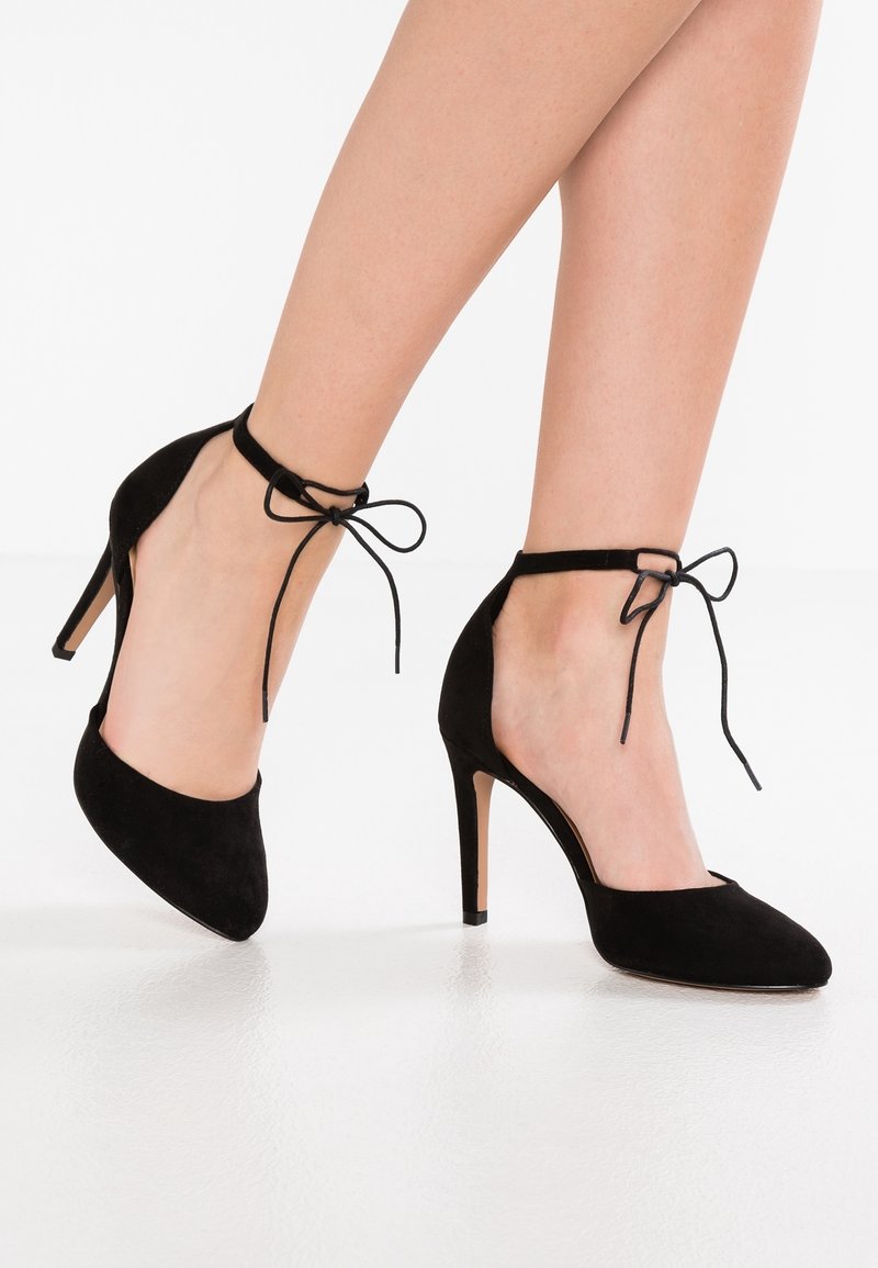 Chaussures à talons hauts en suède noir avec des pointes en aval et des lacets à la cheville. Elles présentent une texture lisse et des talons aiguilles élancés.
