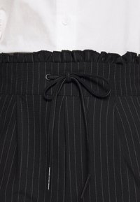 Svart tyg med smala ränder och en volangmidja, utrustad med dragsnöre och veck för en strukturerad silhuett. Vit skjorta med knappar i bakgrunden.