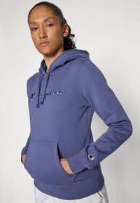 Person med krøllete hår i hestehale iført en lilla Champion hettegenser med hendene i lommen foran, stående mot en enkel lys bakgrunn.