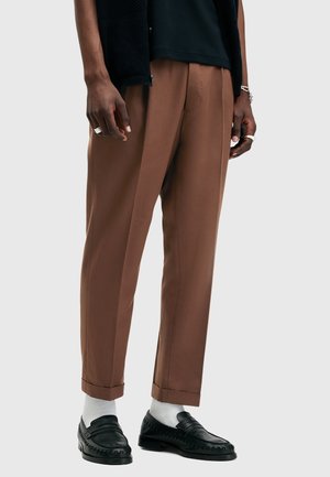 Mężczyzna model ubrany w brązowe, dopasowane spodnie, czarne mokasyny z białymi skarpetkami oraz dodatki na ręce na jasnym, jednolitym tle.