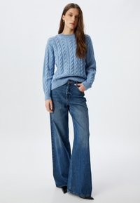 Un maglione azzurro a maglia intrecciata con scollo a rotondo, abbinato a jeans blu a vita alta e a zampa. L'outfit presenta una vestibilità rilassata e una sottile texture.