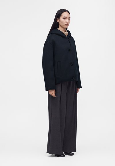 Femme aux longs cheveux noirs portant un manteau noir à capuche boutonné, un pantalon large gris foncé, des chaussures noires et un pull à col roulé beige.