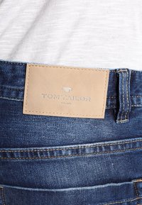 Denimjeans med en slät, mörkblå textur som har en beige läderlapp med texten "TOM TAILOR" på den bakre midjebandet och sydda kanter.