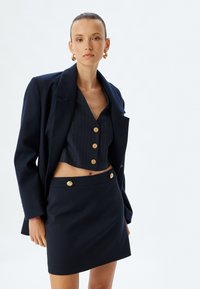 Navy blazer met gegoten revers, gecombineerd met een gestreept cropped top met gouden knopen en een bijpassende hooggetailleerde rok met gouden accenten.