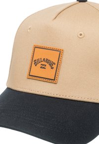 Casquette de baseball beige avec une visière noire, présentée avec une étiquette en cuir marron portant le nom de la marque "Billabong" en relief noir. Tissu texturé.