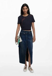 Tamsiai mėlynas trumpas marškinėliai su raudonu grafiniu dizainu, derinami su ilga džinsine sijonu su priekiu iškirpte ir šviesiai pilka delnine su spalvingais akcentais.