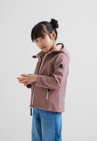 Pink soft-shell jacket with a hood, featuring a front zipper and side pockets. Worn over light blue jeans, with a logo patch on the sleeve.