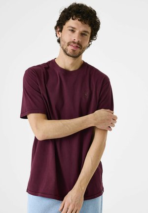 Bordeauxrotes Baumwoll-T-Shirt mit kurzen Ärmeln, Rundhalsausschnitt und kleinem Logodetail auf der Brust. Weiche Textur und lockere Passform.