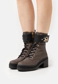 Hnědé vzorované kotníkové boty s černým manžetovým lemem, zlatými detaily, silnou platformou a šněrovacím designem. Zipový detail na boku.