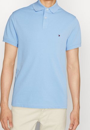 Mand iført lyseblå kortærmet polo med to knapper og lille logo på venstre bryst, stylet med beige bukser.