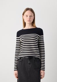 Tengerészkék és törtfehér csíkos pulóver, hajónyakkivágással és hosszú ujjakkal, fekete farmer szoknyával párosítva. Sima textúrájú és laza szabású.